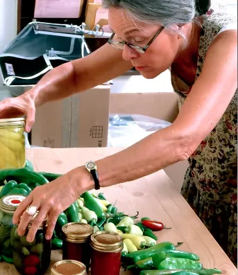 A woman picks up glass jars of canned vegetables from a group of jars surrounded by fresh chili peppers.