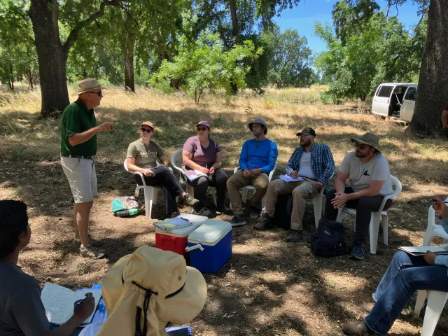 Man in polo and shorts and hat at left speaks with a group of students under shade trees during a farm tour