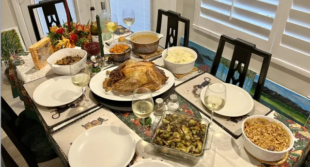 A dining table with stuffing, sweet potatoes, gravy, a roast turkey, mashed potatoes, Brussels sprouts, green bean casserole, and five glasses of wine.