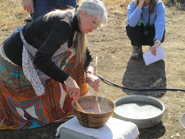 A woman with a spoon in her hand leans over a basket set over a cookstove on what looks like an area of brown grass