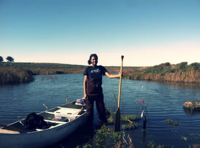A woman stands next to a canoe in the water, holding a paddle upright