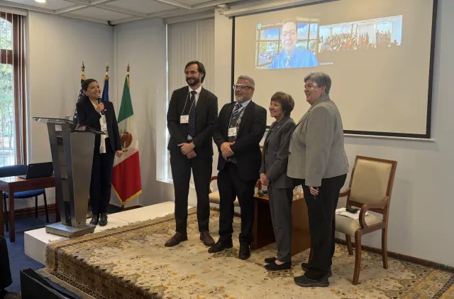 A woman at a lectern speaks into a mic as she looks at four people on stage. On a screen behind them, a man in split screen and large group of people.