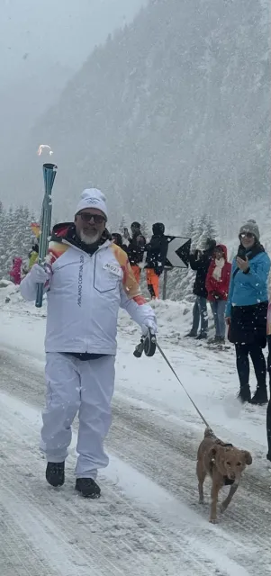 Snow flakes fall as Matteo carries torch in right hand and holds Saba's leash in his left hand as they walk along the snowy path lined with onlookers.