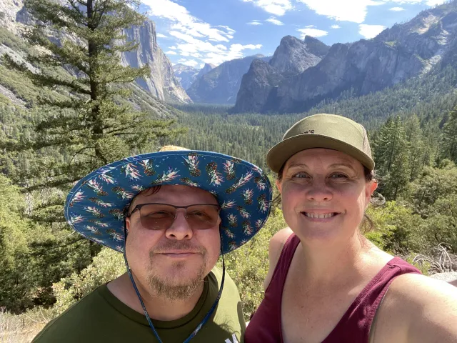 A man and woman pose for a selfie at the Tunnel View vista, with trees and Yosemite Valley behind them