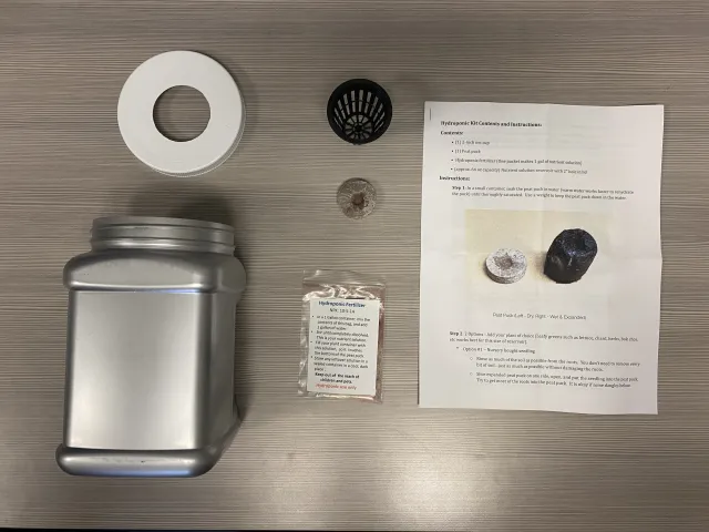 A hydroponic starter kit is laid out on a table, including a container, net cup, growing medium, seeds and printed instructions for setting up a simple system.