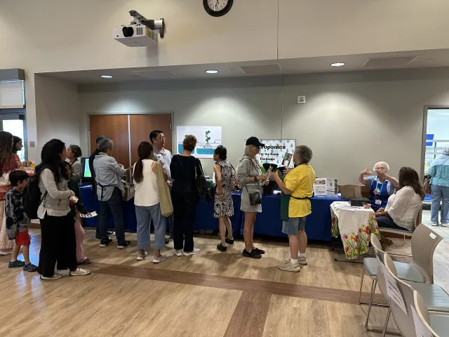 A group of people stand in line at tables inside a community room, speaking with volunteers and viewing hydroponic displays at an educational outreach event.