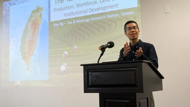 A man in navy blue button-down shirt and glasses gestures while speaking at a podium; his slide behind him features the island of Taiwan