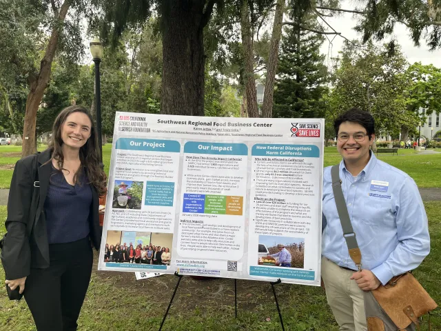 Anne and Nestor stand on opposite sides of a Southwest Regional Food Business Center poster describing the project, its impact and how federal disruptions harm California.
