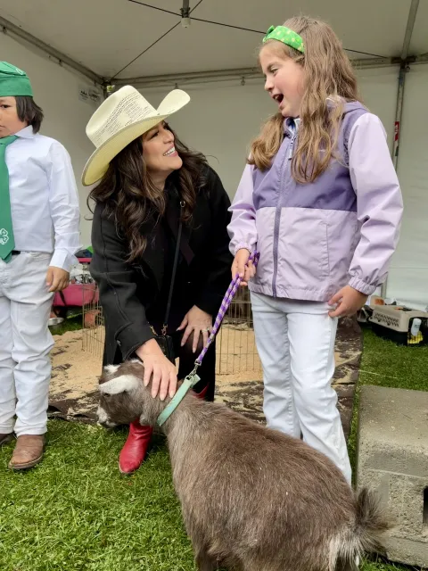 Woman wearing a cowboy hat pets a goat as she listens to a 4-H member who is holding the goat's leash.