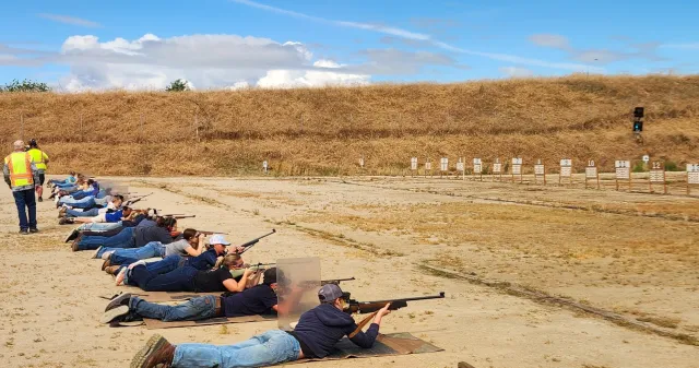 Youth participant lying on their stomach at a rifle range while aiming a rifle at a target during a supervised shooting sports activity.