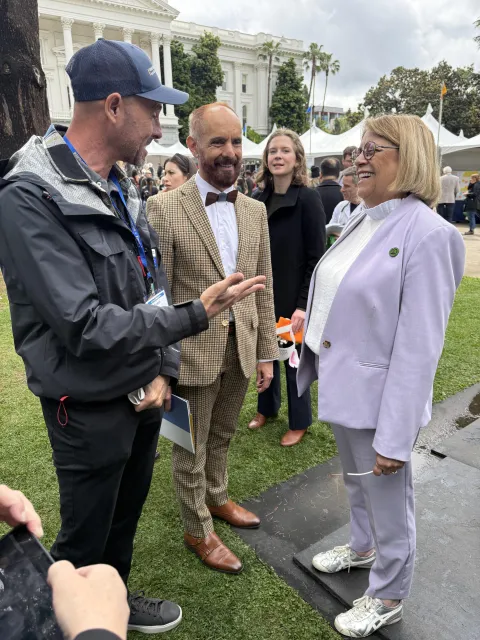 A man on the left gestures with his right hand. The man and woman he's speaking to are laughing.