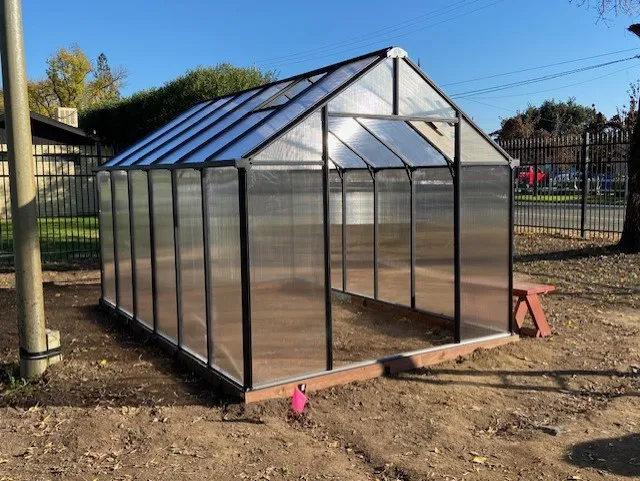 A newly installed greenhouse with a black metal frame and translucent panels, situated on bare soil in a fenced outdoor area. The structure features a peaked roof and an open doorway, with a small red bench in the background.