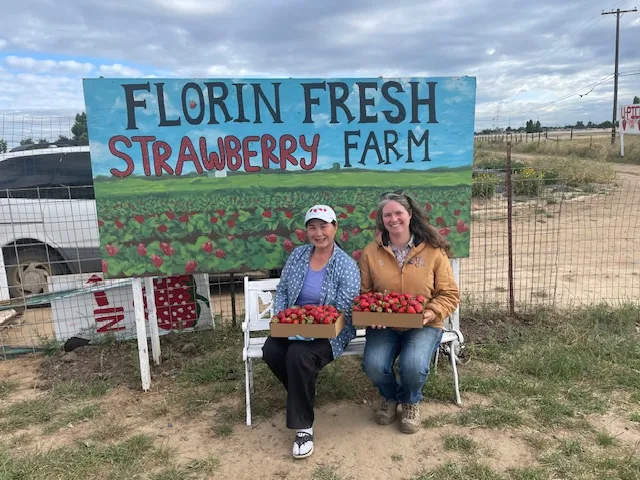 Farmer Muang, left and Margaret Lloyd sit on chairs while holding baskets of strawberries under the Florin Fresh Strawberry Farm sign