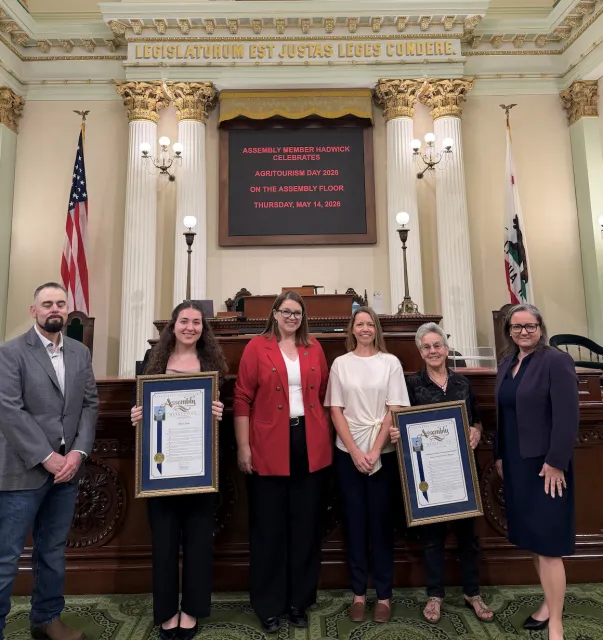 Six people stand in front of U.S. flag in the Assembly chamber. Two of the women are holding framed resolutions.