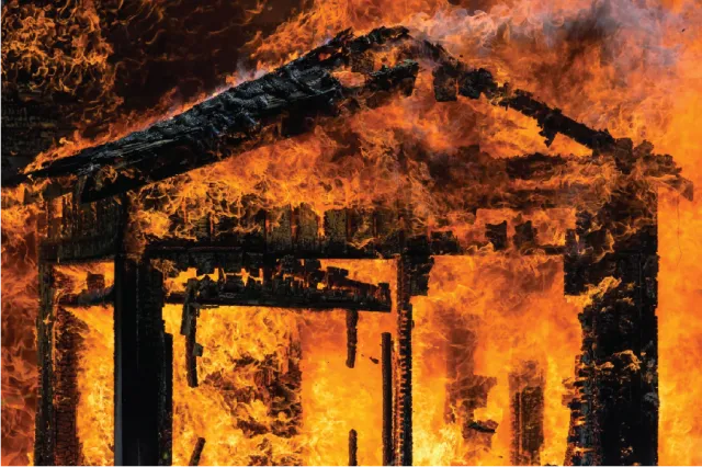 A wooden structure engulfed in intense orange flames, with charred beams and a roof that is about to collapse visible.