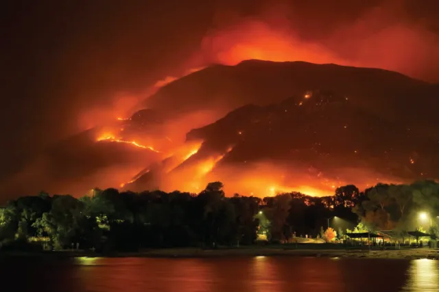 A wildfire rages on a mountainside at night, with flames illuminating the smoke and sky in vibrant oranges and reds, overlooking a dark treeline at the edge of a body of water.