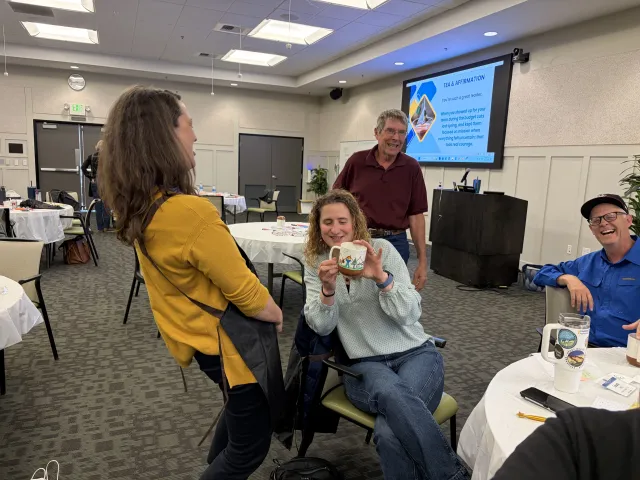 Three people laugh as a woman holds up her decorated mug