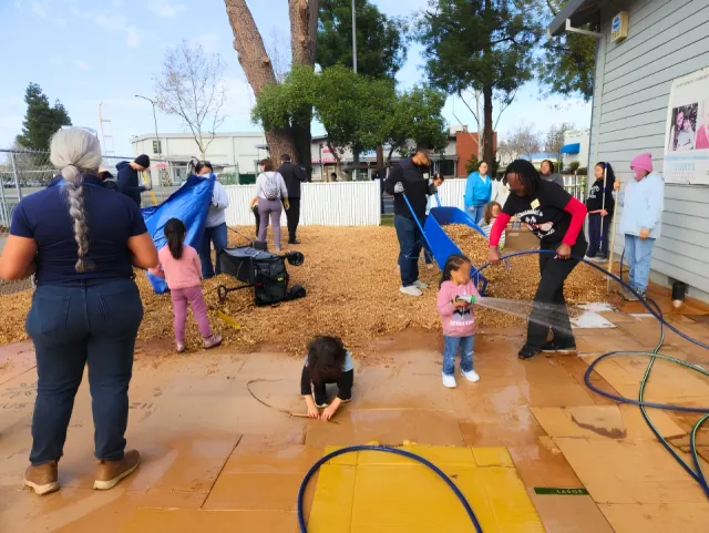 Families working together on an outdoor landscaping project. Participants are spreading wood chips, watering soil and working with wheelbarrows, shovels, and hoses in a neighborhood setting.