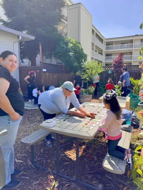 An outdoor gathering in a garden area with families engaging in activities. In the center an adult and child interact at a table with colorful flash cards.
