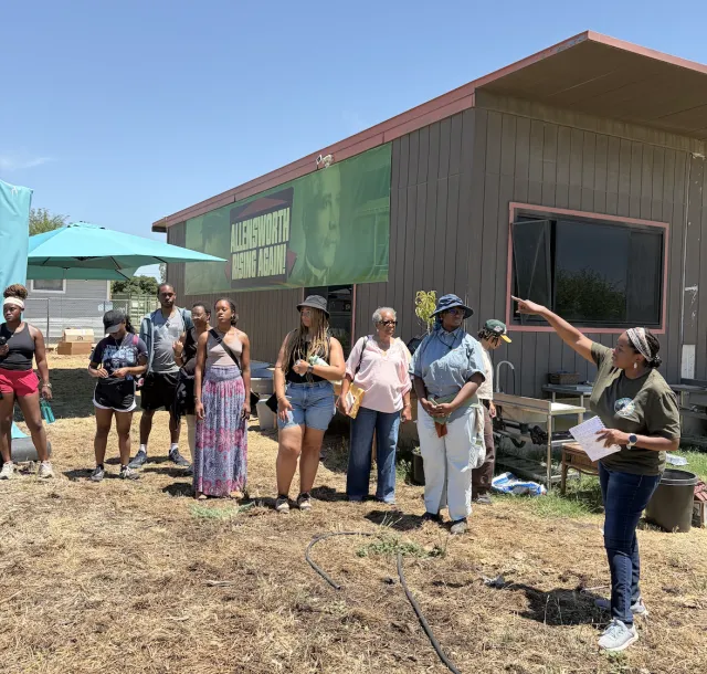 Nine people listen to a woman speak outdoors in front of a building with a banner that reads: Allensworth Rising Again