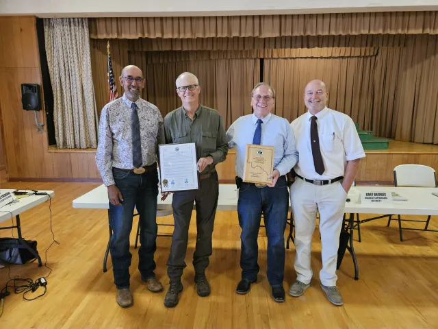 Four men stand in front of a stage in a conference room. David is holding a framed document, another man is holding a plaque with the shape of Lassen County engraved in it.