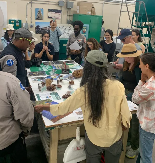 Group of people stand inside around a table and listen to a speaker.