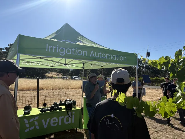 People in a vineyard gather around a man speaking in a tent labeled "Irrigation Automation."