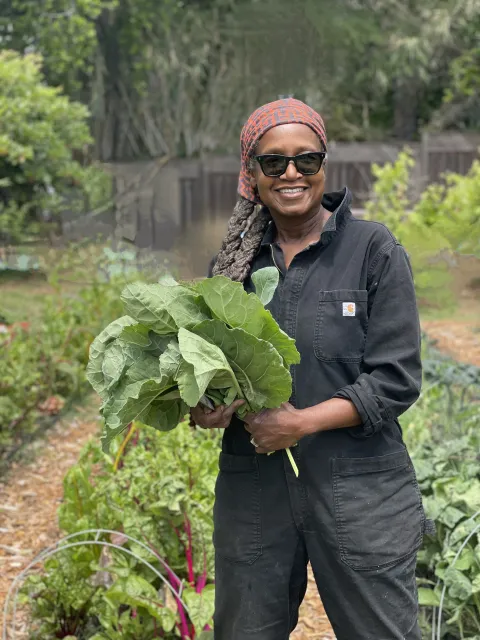 A woman holds a bunch of collard greens while standing in a garden