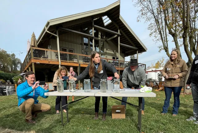Hope, speaking to people gathered around a table, gestures to five glass jars with soil clods floating in water