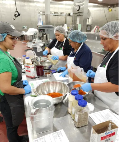 Four people wearing hairnets prepare food on a long steel table in a kitchen