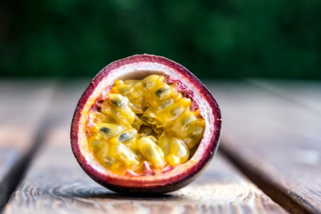 Halved passion fruit with glossy yellow-orange pulp and small seeds, showing the purple-red rind on a rustic wooden surface against a blurred green background.