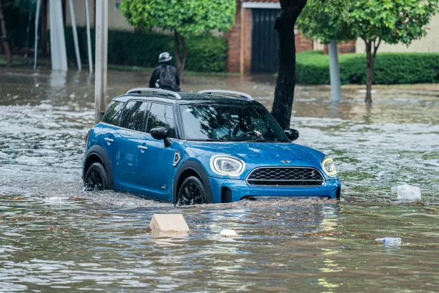 Car drives, partially submerged, on flooded roadway as debris floats by in the high water