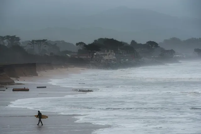 Rain lashes a coastal community under gray skies as a surfer holding a surfboard walks away from the high waves and waterline