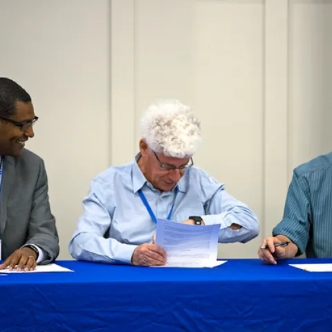 Three men sit at a table signing documents