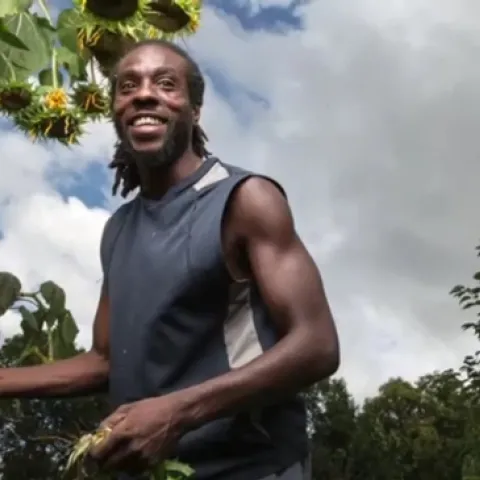 Sacramento farmer Chanowk Yisrael, shown from the waist up holding a shovel handle, stands among crops.