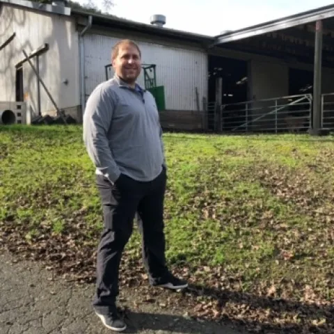 Keith Taylor stands outside on a muddy, grassy area with a building and tree behind him.