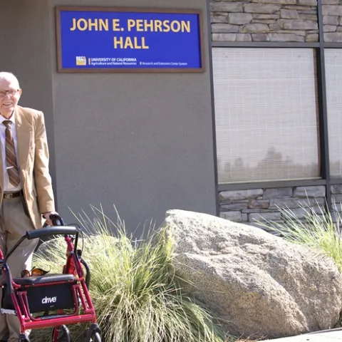 John Pehrson, standing with a walker, poses beside the building sign bearing his name.