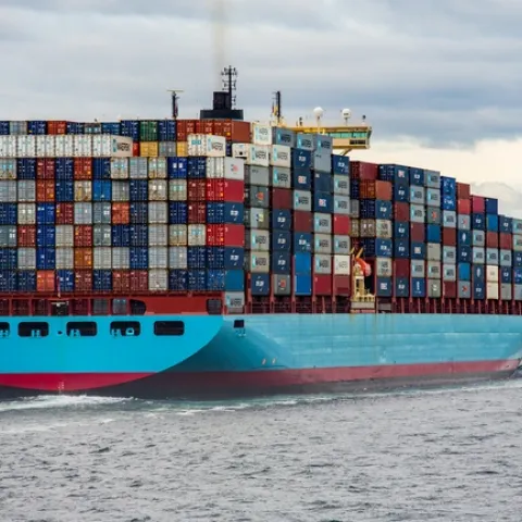 A ship piled high with shipping containers sails out of a port.