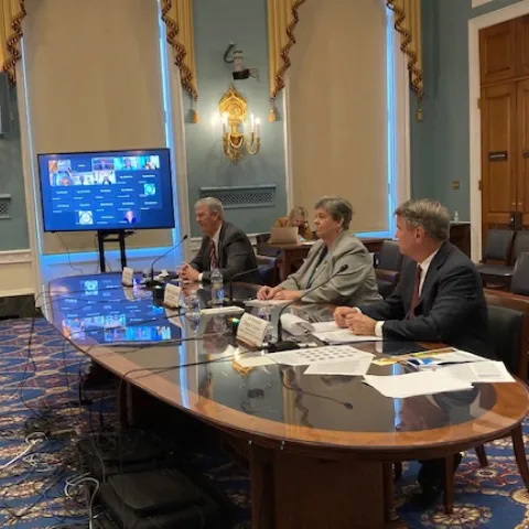 Glenda Humiston sits at a large oval table flanked by two men in dark suits. At one end of the table is a computer screen showing images of people joining the meeting via Zoom.