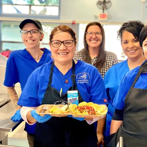 Vanessa Zajfen, wearing a hairnet and surrounded by four colleagues, holds a school lunch tray displaying a meal made with fresh produce.