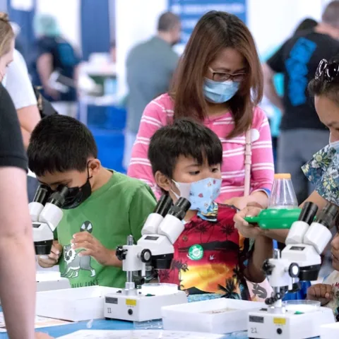 Three dark-haired kids assisted by two women use microscopes.