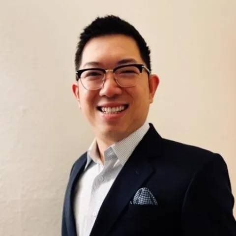 A professional headshot of Cuong Nguyen. He is smiling warmly at the camera, wearing rectangular-framed glasses, a dark navy blazer, a light blue striped dress shirt, and a patterned pocket square. The background is a plain, neutral off-white wall