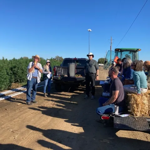 Standing beside a field of hemp, Bob Hutmacher speaks into a mic as growers and others sit on hay bales listening.