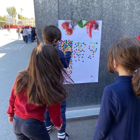 Three students face away from the camera towards a wall with a poster. The poster has images of vegetables, and students place colorful circle stickers in columns under the images for "Love it," "Like it," "Tried it,"