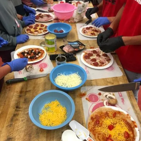 A group of students wear gloves and surround a table laden with small pizzas and pizza toppings.