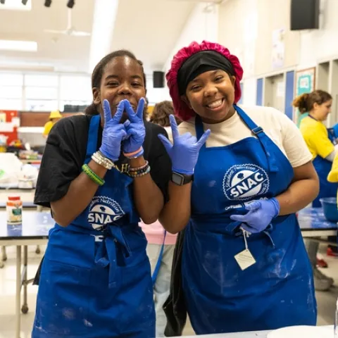 Two girls smiling in a school cafeteria with blue aprons and food prep gloves on.