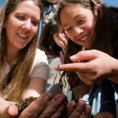 Smiling young girl holding a worm on her finger, while adult holds soil in her hands nearby.