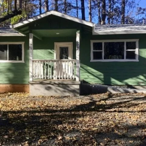 An intact green house with no vegetation in front. Several singed trees behind the house.