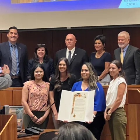 Nine people in two rows posed with a large certificate displayed in the middle. Located in Board chambers
