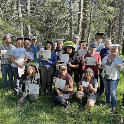 A group of smiling people wearing graduation caps hold up certificates in a grassy, wooded area.